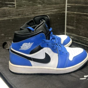 Nike Air Jordan 1 blå och vit US8 - Nike Air Jordan 1 sneakers i blått, vitt och svart med klassisk high-top siluett. Skorna har snörning, perforerad tå och ikoniska Jordan Wings-loggan på sidan. Swoosh i svart på sidorna och blå detaljer på häl och snören. Ovandel i skinn och syntet, insida i textil.
