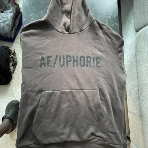 Brun ärmlös hoodieväst AE/UPHORIE - Säljer en brun hoodieväst från AE/UPHORIE med stor magficka och tryckt logga på bröstet. Västen är ärmlös och har en klassisk huva, perfekt för en avslappnad streetstyle-look.
