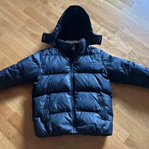 Mörkblå pufferjacka Tommy Hilfiger - Säljer en mörkblå pufferjacka från Tommy Hilfiger med huva och dragkedja. Jackan har två fickor med dragkedja och klassisk quiltad design. Insidan av kragen har snygga ränder i rött, vitt och beige. Perfekt för kalla dagar. Har ett lite jack i bak men inte mycket som syns. Ny pris 3500.