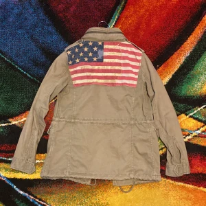 Unik vintage Feildjacket från Ralph Lauren med USA-flagga perfekt skick  - Snygg olivgrön parkajacka från Denim & Supply Ralph Lauren med coolt USA-flagga-tryck på ryggen. Jackan har flera fickor, snörning i midjan och detaljer med dragkedja vid kragen. Perfekt för dig som gillar unika plagg och unika plagg. Storlek S väldigt svåra att hitta skick som ny 10/10 skick Dm för fler bilder eller frågor 🪖🫒⚡️