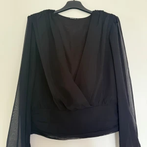 Svart omlottblus med transparenta ärmar - Snygg svart blus från Ginatricot med omlottdesign och v-ringning. Blusen har långa, transparenta ärmar i tunn polyester som ger en cool och luftig känsla. 