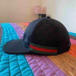 Svart Gucci keps med röd/grön rand - Snygg svart keps från Gucci med diskret GG-mönster över hela och en klassisk röd/grön rand på sidan. Kepsen har böjd skärm och justerbar rem bak. Perfekt för dig som vill ha en lyxig och trendig accessoar. Helt ny och bästa kopian