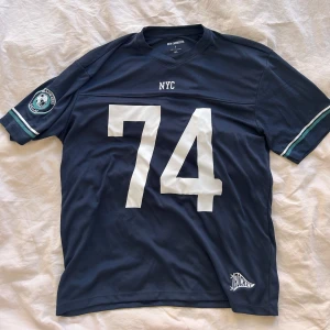 Mörkblå NYC t-shirt med tryck - Sportig mörkblå t-shirt med stort vitt tryck '74' på framsidan och NYC-text ovanför. V-ringad hals och korta ärmar med vita och gröna ränder. Patch på ena ärmen och Hockey-logga nedtill. Perfekt för en chill streetstyle. Oversized