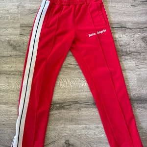 Palm Angels röda trackpants M - Palm Angels röda trackpants med vita och svarta ränder längs sidorna. Byxorna har elastisk midja, sidofickor och dragkedja vid benslut. Materialet är 100% polyester och loggan är tryckt på vänster lår. Snygg streetwear-vibe och perfekt för en chill look.