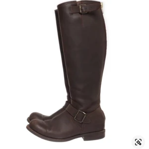 Bruna stövlar i skinn med spänne - Snygga bruna stövlar i äkta skinn. De har rund tå, platt sula och detaljer med spännen upptill och vid ankeln. Dragkedja baktill gör dem enkla att ta på. Perfekta för dig som gillar klassisk stil och vill ha boots som funkar till ridning.