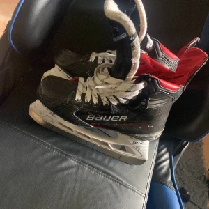 Bauer Vapor X4 hockeyskridskor - Säljer ett par Bauer Vapor X4 hockeyskridskor med svart och röd design, vita snören och robust plastskena. Insidan är fodrad för extra komfort och stöd. Perfekt för dig som vill ha grym kontroll och fart på isen. Snygg sportig look och slitstarka material.