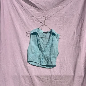 Ljusblå ärmlös linnetopp från Mirrou - Superfin ljusblå linnetopp från Mirrou i 100% bomull. Toppen är croppad, har krage, djup v-ringning och knappar framtill. Perfekt för varma dagar och enkel att styla med jeans eller kjol.