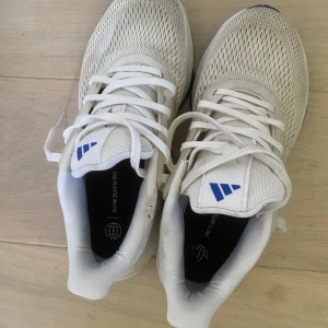 Vita Adidas sneakers med blå sula - Fräscha vita Adidas sneakers med blå detaljer och sula. Ovandelen är i ventilerande mesh och har klassiska snörningar. Snyggt Adidas-märke på plösen och diskreta ränder på sidan. Perfekta för träning eller chill vardagsstil. Lite skitiga men går nog bort om man tvättar noggrant.