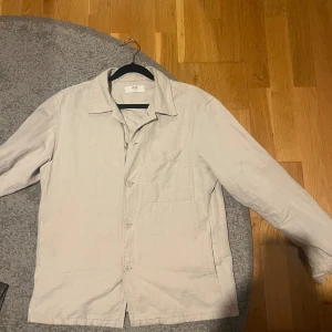 Beige overshirt från Uniqlo  - Snygg beige overshirt från Uniqlo. Skjortan har klassisk krage, bröstficka och knäppning framtill med tre knappar. Tillverkad i ett lätt och luftigt linnematerial, perfekt för varmare dagar. Passar dig som gillar enkel och stilren design. Stl XS på overshirten men passar perfekt på som S! Ej använd är som ny! Se bild 2 för att se tydligare färgen 