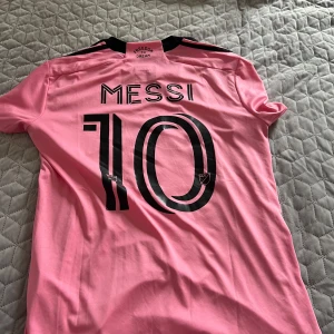 Inter Miami Messi rosa matchtröja - Snygg Inter Miami fotbollströja i rosa med svarta detaljer, Messi och nummer 10 på ryggen. Adidas-logga och klubbmärke på bröstet, samt MLS-märke på ärmen. Tillverkad i lätt och ventilerande material, perfekt för träning eller match.
