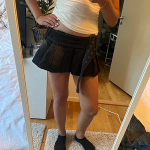 Svart plisserad minikjol med bälte - Supersnygg svart plisserad minikjol i jeansmaterial med bred midja och coolt bälte med metallspänne. Kjolen har en loose passform och ger en edgy vibe med sitt korta snitt och detaljerade midjeband. Perfekt för dig som vill sticka ut med en trendig look.