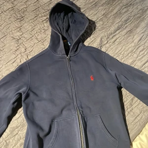 Mörkblå hoodie från Polo Ralph Lauren - Säljer en mörkblå hoodie från Polo Ralph Lauren med röd logobrodyr på bröstet. Tröjan har huva, dragkedja framtill och två fickor. Insidan är mjuk och fleecefodrad, perfekt för kyliga dagar. Klassisk och stilren design som funkar till jeans eller joggers. Passar om man e 155-160 cm. Den ser lite missfärgad ut men den har en jätte fin blå färg som har Holly jätte bra