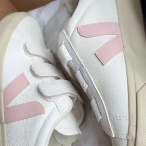 Vita sneakers med rosa detaljer - Snygga vita sneakers från Veja med ljusrosa V-logga på sidan och praktiska kardborreband. Skorna har en rund tå och är tillverkade i skinn med beige sula. Perfekta för dig som gillar stilrena och fräscha sneakers med en soft touch av rosa. Perfekta nu till hösten hör av er om ni vill ha fler bilder elelr diskutera nått skorna är använda nån gång💗💗skorna är köpta på vejas egna hemsida och köpta för 1750kr