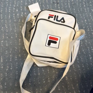 Vit axelväska från FILA - Snygg vit axelväska från FILA med klassisk logga i svart och rött på framsidan. Väskan har en fyrkantig form, svart kant och justerbar axelrem. Perfekt storlek för mobil, plånbok och nycklar. Stängs med dragkedja och har en extra ficka framtill.