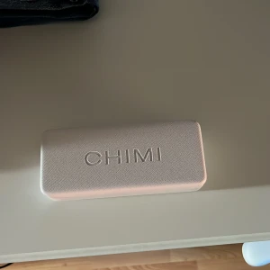 Grå CHIMI solglasögon med fodral - Snygga solglasögon från CHIMI med transparent grå båge och svarta glas. Modellen har en fyrkantig form som ger en cool vibe. Medföljer ett stilrent vitt fodral och en mjuk tygpåse för förvaring. Perfekt accessoar för soliga dagar.