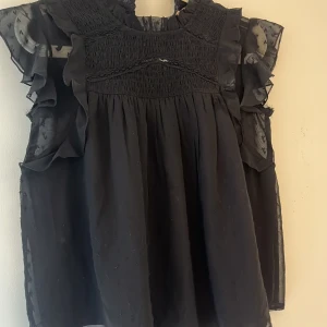 Svart blus med volang från Gina Tricot - Supersnygg svart blus från Gina Tricot med transparenta volangärmar och smock upptill. Blusen har en lätt rynkad struktur och små broderade detaljer på tyget. Perfekt för dig som gillar romantiska vibes och vill ha något extra till din outfit. Man kan också du upp den om den är för lång men jag tycker den är perfekt 😍 