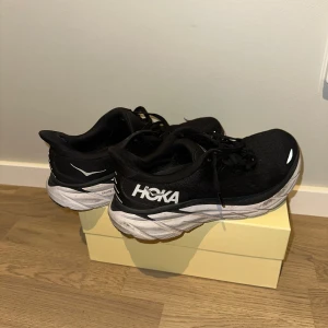 Svarta Hoka Clifton 8 sneakers - Säljer ett par svarta Hoka Clifton 8 sneakers med vit sula och coola detaljer i vitt. Är lite slitna se bild. Men fungerar bra annars 