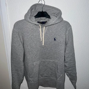 Grå hoodie från Polo Ralph Lauren - Säljer en grå hoodie från Polo Ralph Lauren med klassisk broderad logga på bröstet. Tröjan har huva med vit snörning, känguruficka och långa ärmar. Mjuk bomullskvalitet som är perfekt för chill dagar. Enkel och stilren design som funkar till allt. Pris kan diskuteras
