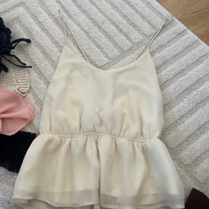 Ljusbeige linnetopp med snörning - Supersöt ljusbeige linnetopp med tunna axelband och volang nedtill. Toppen har snygg snörning i ryggen som ger en extra detalj. Perfekt för varma dagar och enkel att styla med jeans eller kjol.