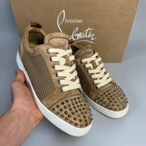 Beige sneakers från Christian Louboutin - Snygga beige sneakers från Christian Louboutin i mocka med coola silverfärgade nitar på tån. Skorna har vita snören, vit sula och den klassiska röda undersulan. Sidorna har ett diskret mönster och materialet känns lyxigt och mjukt.