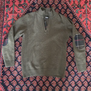 Olivgrön Barbour stickad tröja half zip - Snygg olivgrön stickad tröja från Barbour med half zip och hög krage. Tröjan har ribbade muddar, rutiga förstärkningar på armbågarna och diskreta detaljer i rött och vitt. Perfekt för dig som gillar klassisk stil med en twist.
