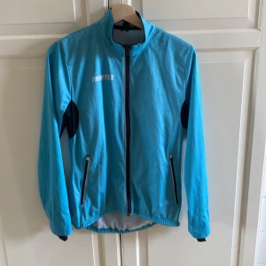 Blå vindjacka från Trimtex - Sportig blå vindjacka från Trimtex med svarta detaljer under armarna och dragkedjor på fickorna. Jackan har hel dragkedja framtill, hög krage och elastisk nederkant. Perfekt för orientering, om man inte vill lägga så mycket pengar på nya kläder. Storlek M. Skicket är helt okej, finns lite små revor på jackan men inga hål och inget som påverkar jackan. Köpte den begagnad på Tradera, men säljaren har inte uppgett skicket. 