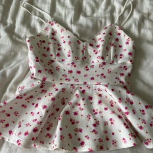 Blommig peplumtop från Gina Tricot - Supersöt vit peplumtop från Gina Tricot med små rosa blommor över hela tyget. Toppen har smala axelband, markerad midja och en utsvängd nederdel som ger en feminin siluett. Perfekt för vår och sommar när du vill ha en romantisk look.