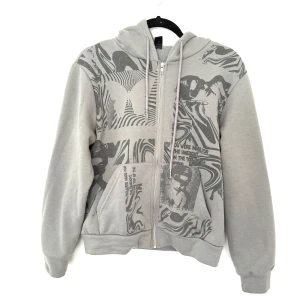 Grå hoodie med grafiskt tryck - Grå hoodie från SHEIN i storlek XS med coolt grafiskt tryck framtill i mörkgrått. Tröjan har huva med snörning, dragkedja och kängurufickor. Långa ärmar, perfekt för en chill och avslappnad stil. Lite nopprig kolla fjärde bilden 