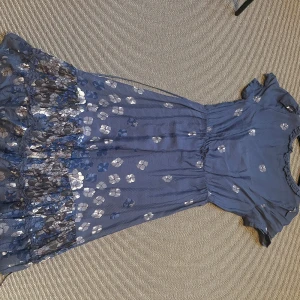 Blommig blå klänning Noa Noa XS - Supersöt långklänning från Noa Noa i blått med silvriga blommor, volangkant nertill och v-ringning med knappar och knytband i midjan. Lätt och luftig, perfekt för sommarens alla tillfällen. Kortärmad och med fina detaljer längs urringningen.