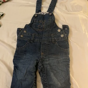 Jeansbyxdress med broderi från H&M ( barn storlek) - Mörkblå jeansbyxdress från H&M i storlek 62. Byxdressen har justerbara axelband, flera fickor och snygga broderade detaljer på bröstfickan och bakfickorna. Tillverkad i bomull med klassisk denimlook och dekorativa knappar i sidorna.