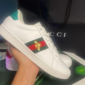 Gucci ace - Snygga vita sneakers från Gucci i skinn med klassiska gröna och röda ränder på sidan och en broderad guldfärgad bi. Skon har snörning, platt sula och metallic-detaljer i rött och grönt på hälen. Perfekt för dig som vill sticka ut med lyxig streetstyle. !! Dem är använda 1 gång inomhus !!