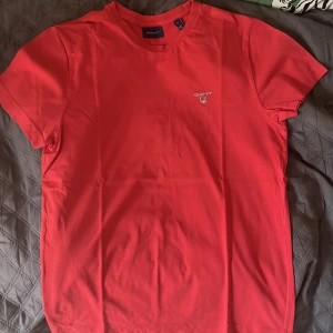 GANT T-SHIRT - RÖD/ROSA - STORLEK S - Snygg röd t-shirt från GANT i mjuk bomull. Klassisk passform med rund hals och korta ärmar. Liten GANT-logga broderad på bröstet. Perfekt för en clean och stilren look. Äkta vara köpt ifrån Boozt.
