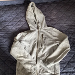 Grå hoodie från Sail Racing med dragkedja - Snygg ljusgrå hoodie från Sail Racing med hel dragkedja framtill och extra ficka med dragkedja på bröstet. Tröjan har huva med snörning och är tillverkad i mjuk bomullsmix. Perfekt för chill dagar eller när du vill ha en sportig look. Dragkedjan På fickan vid bröd är sönder.