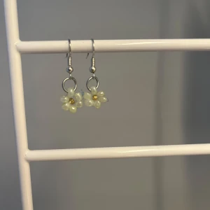Söta blommiga örhängen i silverfärg - Ett par hängande örhängen med små blommor i pärlemorvit och gul detalj i mitten. Örhängena har silverfärgade krokar och ringar, och blommorna är formade av pärlliknande material. Perfekta för att ge en gullig touch till din stil. Det är handgjorda!