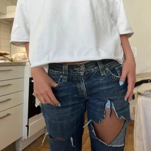 Mörkblå slitna bootcut jeans - Säljer ett par mörkblå jeans med stora slitningar över knäna och bootcut-ben. Jeansen har låg midja och klassiska fickor både fram och bak. Perfekta för dig som gillar en edgy och avslappnad stil.