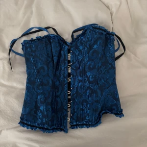 Blå korsett med snörning och mönster - Säljer en blå korsett med snyggt barockinspirerat mönster och svarta samt blå band. Toppen har snörning i ryggen och volangkant upptill och nedtill. Perfekt till evenemang med klädkod. Använt en gång