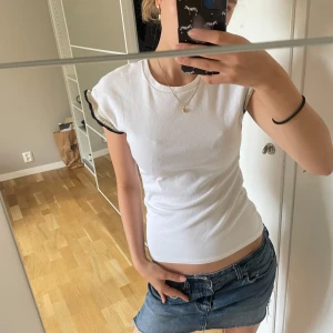 Vit topp med volangärmar och guldtråd - Supersöt vit topp med korta volangärmar som har detaljer i guldtråd och svart kant. Toppen är figurnära och har en enkel, clean look med en twist tack vare de glittriga ärmarna. Perfekt för dig som gillar att sticka ut lite extra.