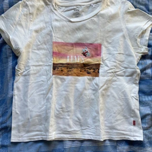 Vit Levi's t-shirt med tryck - Vit t-shirt från Levi's med coolt tryck framtill i rosa och gula toner som föreställer ett landskap och en liten figur. Klassisk passform och rund halsringning, perfekt för en avslappnad stil.