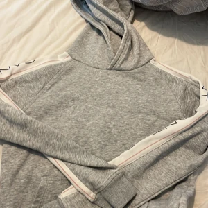 Grå hoodie från GANT med logga - Säljer en grå hoodie från GANT med vita band längs ärmarna där GANT-loggan är tryckt i svart. Tröjan har huva, känguruficka och ribbade muddar. Perfekt för chill dagar och snygg till jeans eller mjukisbyxor.