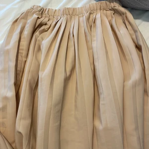 Plisserad beige långkjol - Säljer en superfin plisserad långkjol i beige med bred resår i midjan. Kjolen har ett flowigt fall och passar perfekt till sneakers eller boots. Enkel att styla och ger en chill men ändå classy vibe.