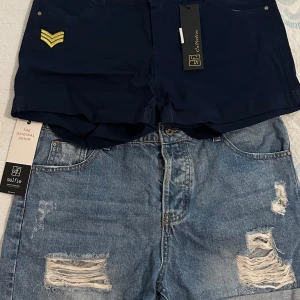 Två par shorts: jeans och marinblå - Säljer två par shorts! Ett par klassiska blå jeansshorts från Selfie med slitna detaljer och upprullade benslut. Det andra paret är marinblå från SF Collection, med en gul broderad detalj framtill och stilren design. Perfekta för varma dagar och snygga till sneakers.
