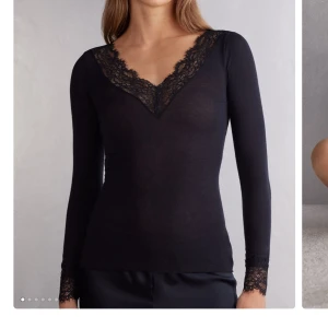 Svart långärmad spetsblus - Supersnygg svart långärmad blus med djup v-ringning och breda spetsdetaljer vid halsringning och ärmslut. Toppens material är tunt och stretchigt, vilket ger en slim passform. Perfekt för dig som gillar en feminin och stilren look. Aldrig använd men prislappen är inte kvar