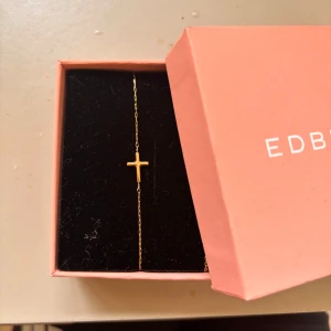 Guldigt armband med kors från Edblad - Snyggt och stilrent armband från Edblad i guldton med en tunn kedja och ett litet kors som detalj. Perfekt för dig som gillar minimalistiska smycken med en personlig touch. Levereras i original ask.