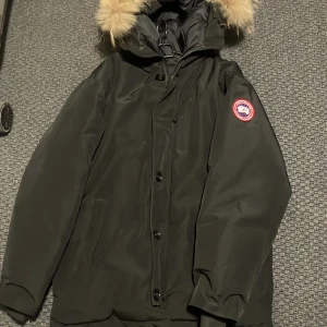 Svart dunjacka från Canada Goose - Säljer en svart dunjacka från Canada Goose med klassisk patch på ärmen och stor huva med fluffig päls. Jackan har dragkedja, knappar och flera fickor. Perfekt för kalla dagar och riktigt snygg streetstil.