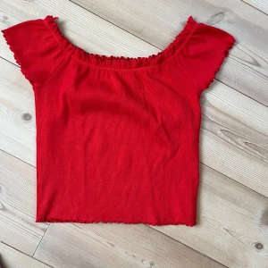 Röd offshoulder topp Gina Tricot - Superfin röd offshoulder topp från Gina Tricot med volangkant vid halsringningen och ärmslut. Toppen är ribbad och har en croppad passform som ger en trendig vibe. Perfekt för att lyfta vilken outfit som helst!