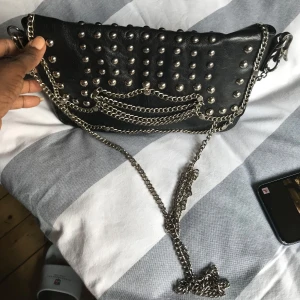 Svart axelväska med nitar och kedjor - Cool svart axelväska i skinn med massor av silvriga nitar och kedjor framtill. Väskan har ett rockigt uttryck och stängs med dragkedja. Perfekt för dig som gillar edgy detaljer och vill sticka ut.