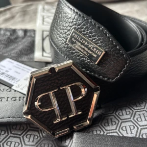 Svart skärp från Philipp Plein i skinn - Exklusivt svart skärp från Philipp Plein i äkta skinn med grov textur. Snyggt silverfärgat PP-spänne i hexagonform och metallbricka med logga. Kommer med originalpåse och etiketter. Perfekt för dig som gillar statement-accessoarer.