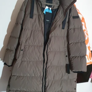 Brun lång pufferjacka Rockandblue - Säljer en lång, brun pufferjacka från Rockandblue i modellen Camilla Coat. Jackan har quiltad design, dragkedja framtill, huva och praktiska fickor med knapp. Tillverkad i återvunnen polyester och dun, perfekt för kalla vinterdagar. Färgen är mole brown.