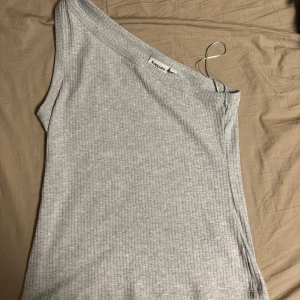 Ljusgrå ribbad oneshoulder topp Kappahl - Snygg ljusgrå ribbad topp från Kappahl med oneshoulder-design. Toppen är ärmlös och har en tight passform som framhäver axlarna. Perfekt för dig som gillar stilrena och trendiga plagg med en twist.