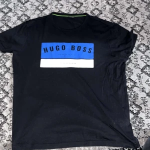 Svart Hugo Boss t-shirt - Svart t-shirt från Hugo Boss med ett stort blått och vitt tryck på bröstet där det står 'Hugo Boss'. Klassisk rund halsringning och korta ärmar. Perfekt för dig som gillar stilrena märkesplagg med tydlig logga.